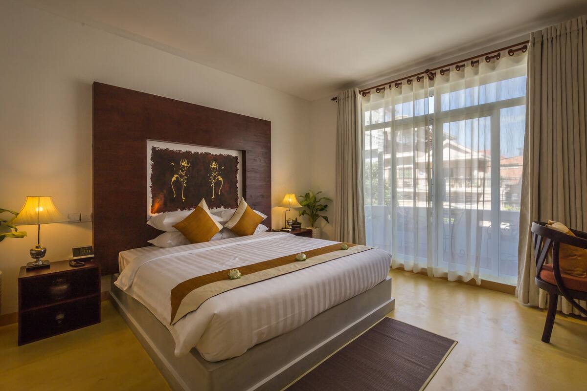 A hotel room with a large bed, two bedside tables with lamps, a nightstand, a window with curtains, and a view of buildings outside.