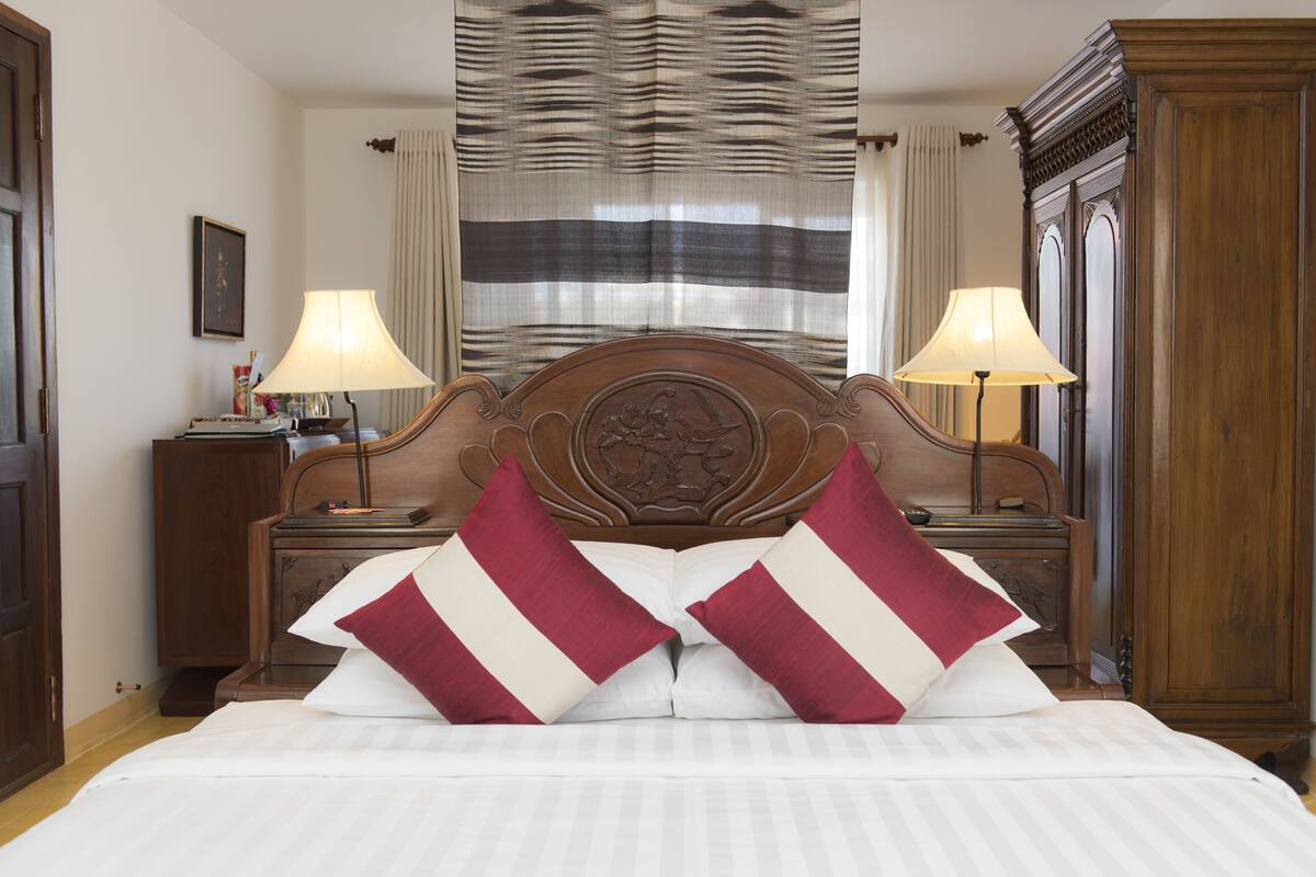 A neatly made bed with two pillows, a wooden headboard, two bedside tables with lamps, and a window with curtains.