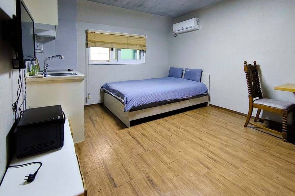 A bedroom with a bed, a desk with a computer, a chair, a window with blinds, and an air conditioning unit.