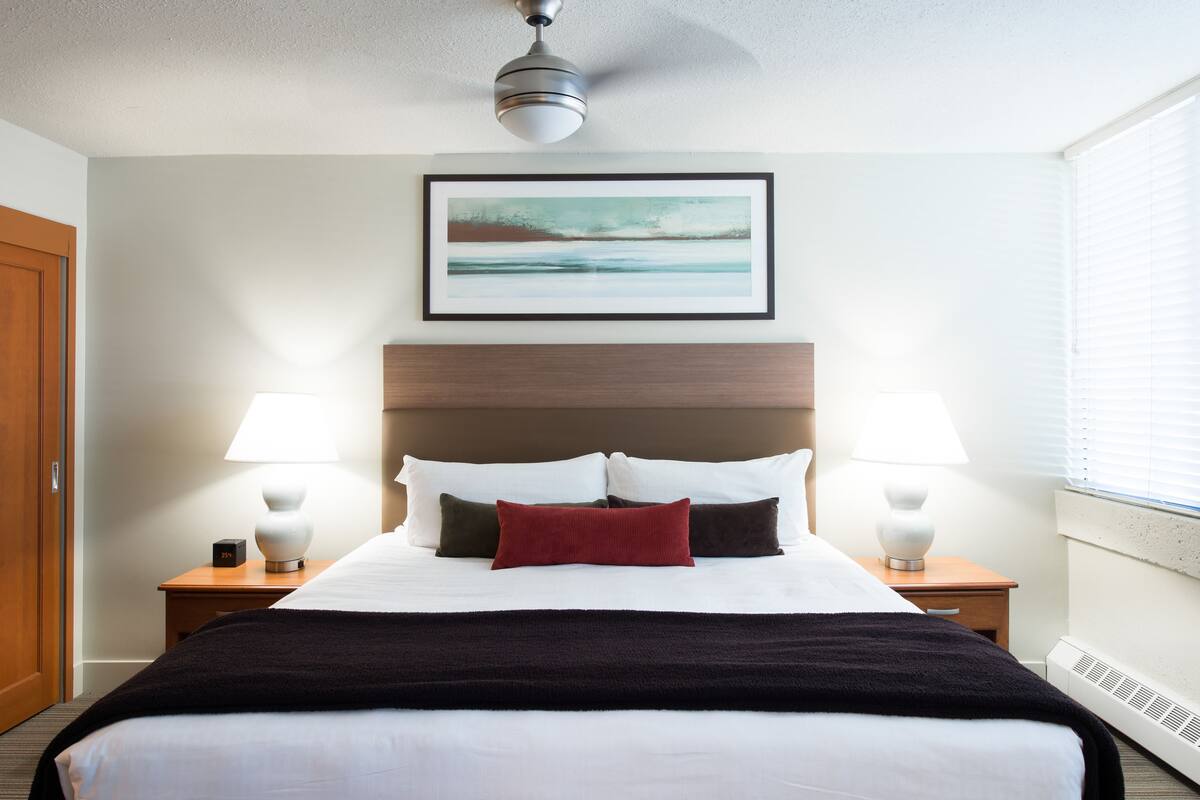 Ein Hotelzimmer mit einem großen Bett, zwei Nachttischlampen, einem Deckenventilator, einem gerahmten Kunstwerk über dem Bett und einem Fenster mit Jalousien.