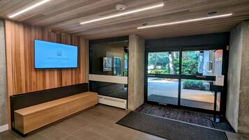 Zona de recepción con un banco de madera, un televisor de pantalla plana mostrando 'West Coast Suites' y una puerta de cristal que da al exterior.