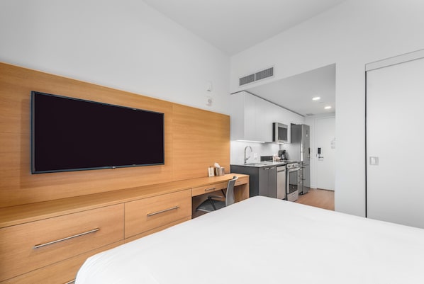 Una habitación de hotel moderna con una cama grande, cabecera de madera, televisor de pantalla plana, escritorio con silla, kitchenette y baño.