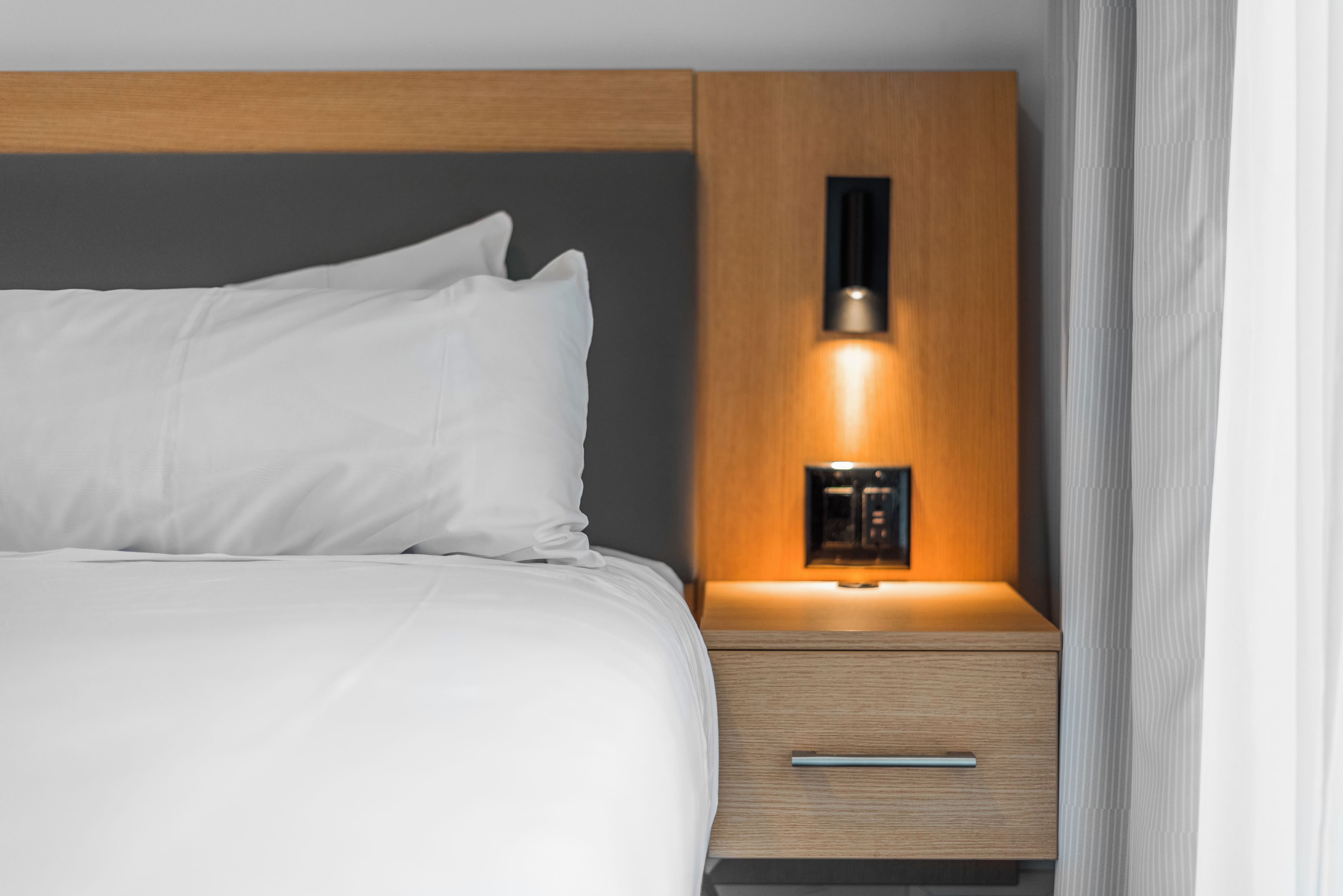 Una cama bien hecha con sábanas blancas y una mesita de noche de madera con cajón y una lámpara fijada a la pared.
