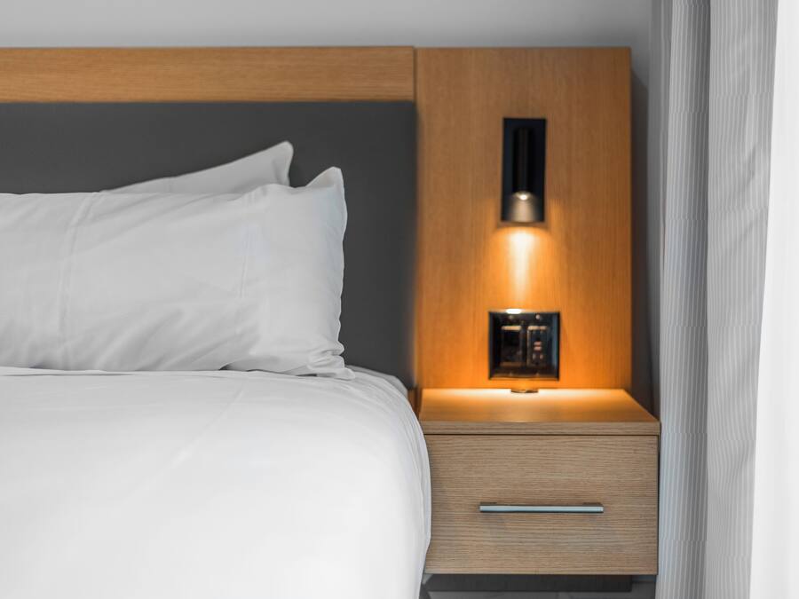 Una cama bien hecha con sábanas blancas y una mesita de noche de madera con cajón y una lámpara fijada a la pared.