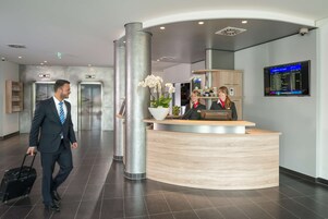 Um saguão de hotel moderno com balcão de recepção, elevadores e um painel de informações de voos digital.