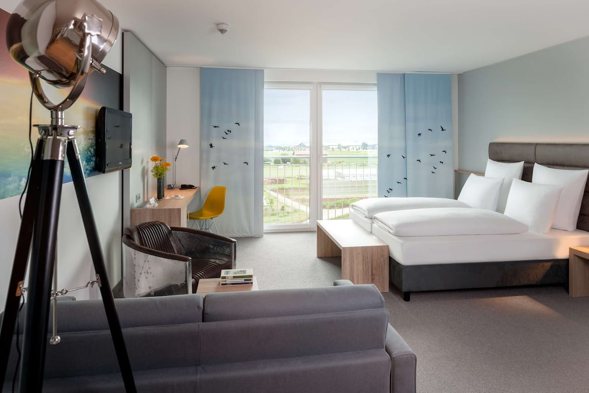 Habitación de hotel con cama, escritorio, silla y un amplio ventanal con vistas al exterior.