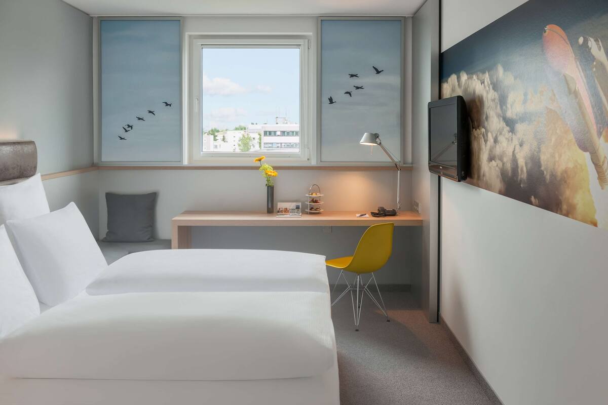 Habitación de hotel con cama, escritorio, silla, ventana y televisor instalado en la pared.