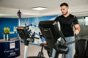 Ein Mann benutzt ein Trainingsgerät in einem Fitnessraum eines Hotels, mit blauer Wand und einem Poster von Fallschirmspringern.