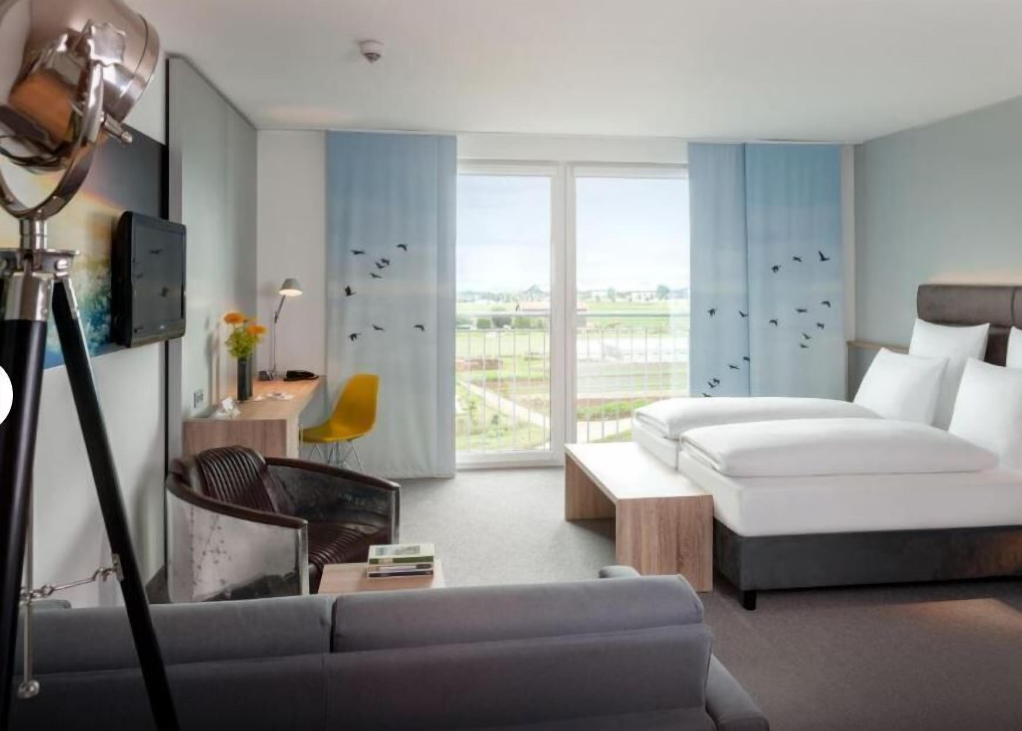 Una habitación de hotel moderna con una cama grande, un escritorio con lámpara, una silla y vistas al exterior.
