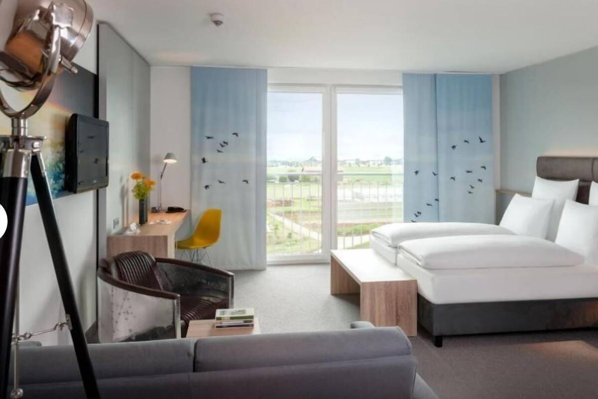 Una habitación de hotel moderna con una cama grande, un escritorio con lámpara, una silla y vistas al exterior.