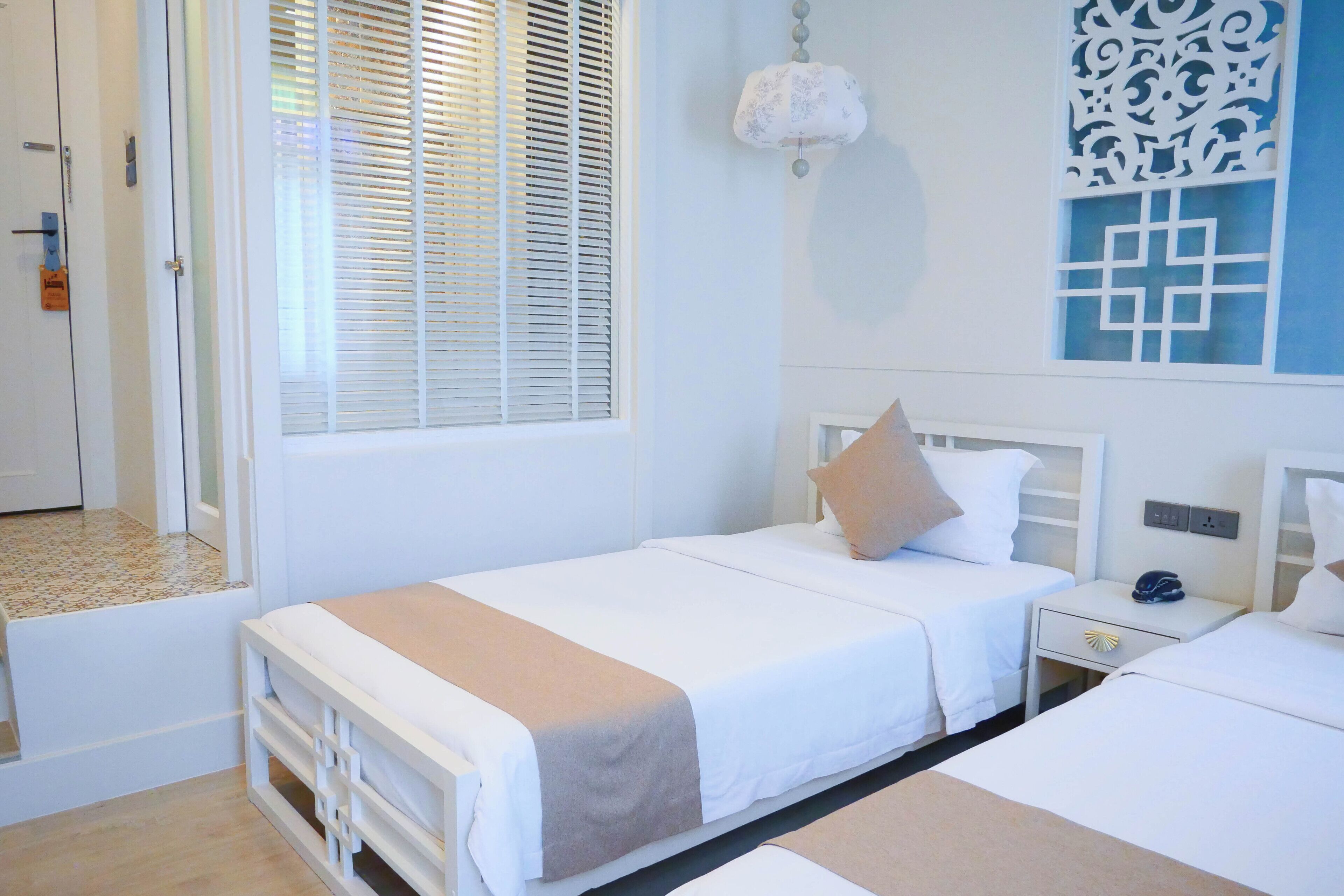 A hotel room with two beds, a window with blinds, a decorative wall panel, and a nightstand with a phone and a decorative item.