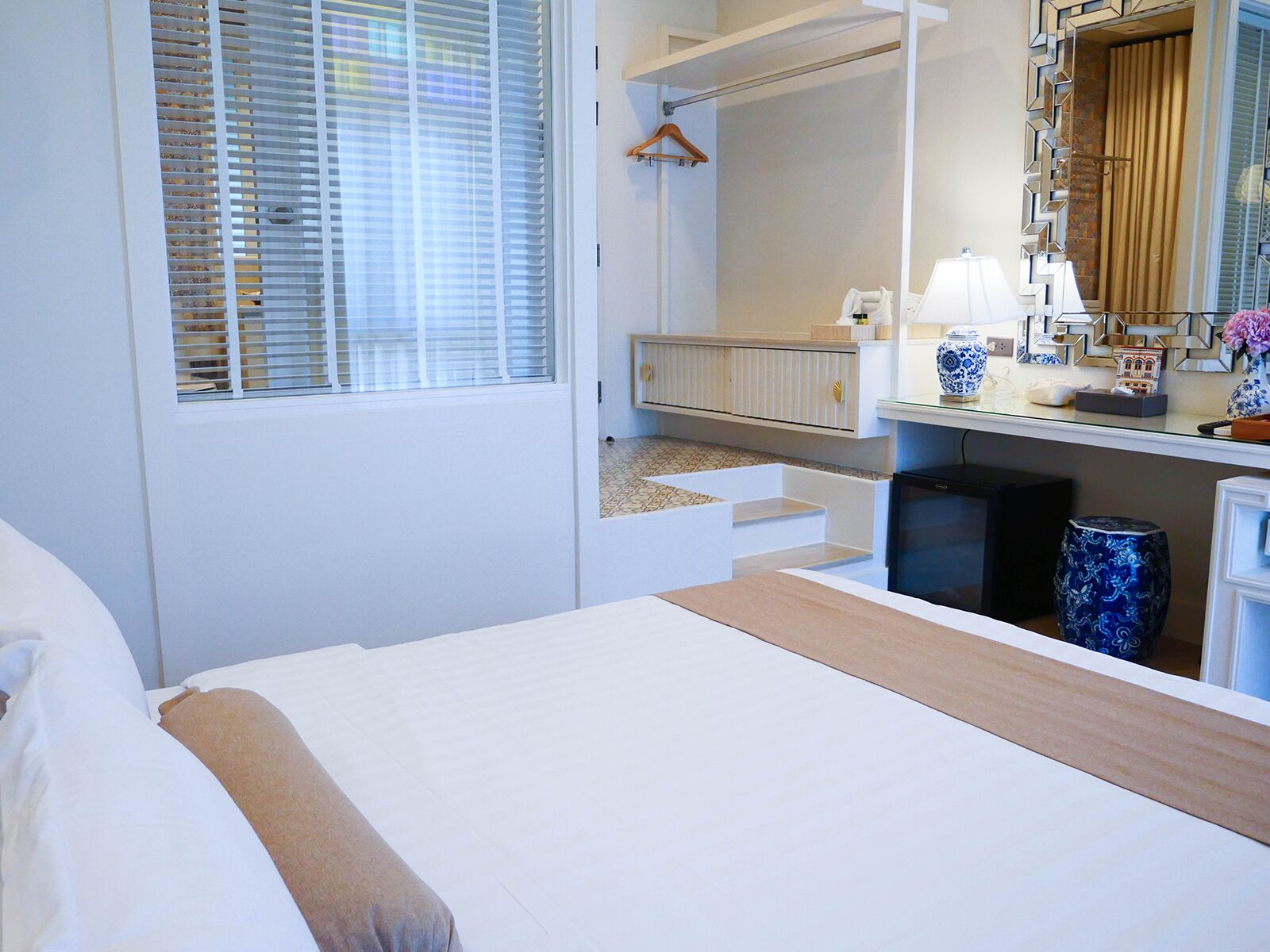Hotellihuone, jossa on sänky, työpöytä lampun kanssa, peili ja ikkuna, jossa on sälekaihtimet.