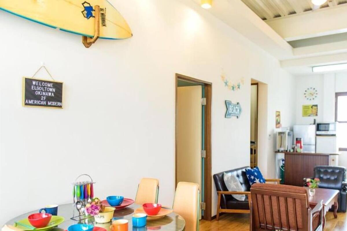 A modern living room with a dining area, a surfboard, and a welcome sign.