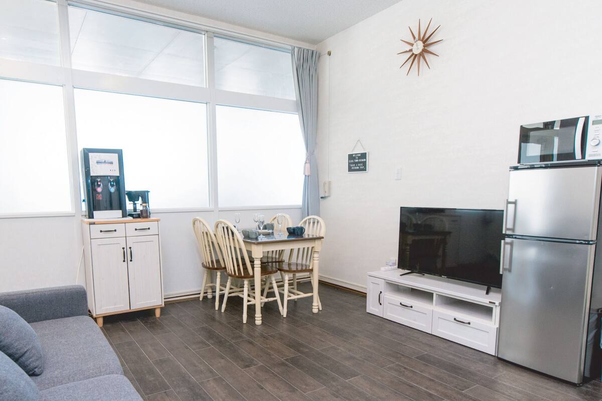 A modern kitchen with a dining area, a grey sofa, and a TV.
