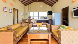 Un salón espacioso con techo alto, una mesa central con superficie de vidrio, mobiliario de mimbre y una cocina al fondo.