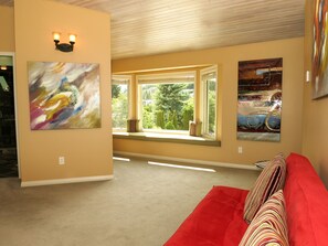 Un salon avec un canapé rouge, deux fenêtres équipées de stores et deux tableaux abstraits accrochés aux murs.
