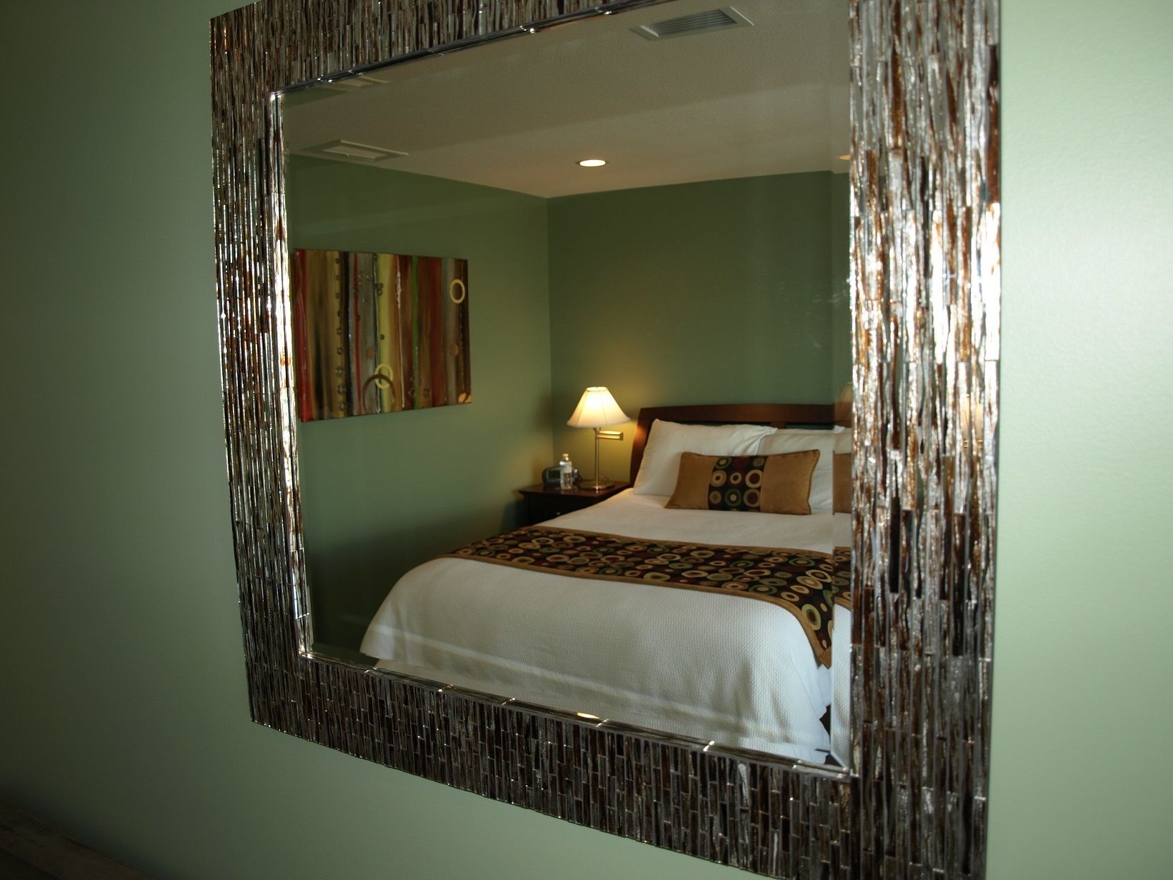 A hotel room with a large mirror, a bed with white bedding, a nightstand with a lamp, and a colorful abstract painting on the wall.