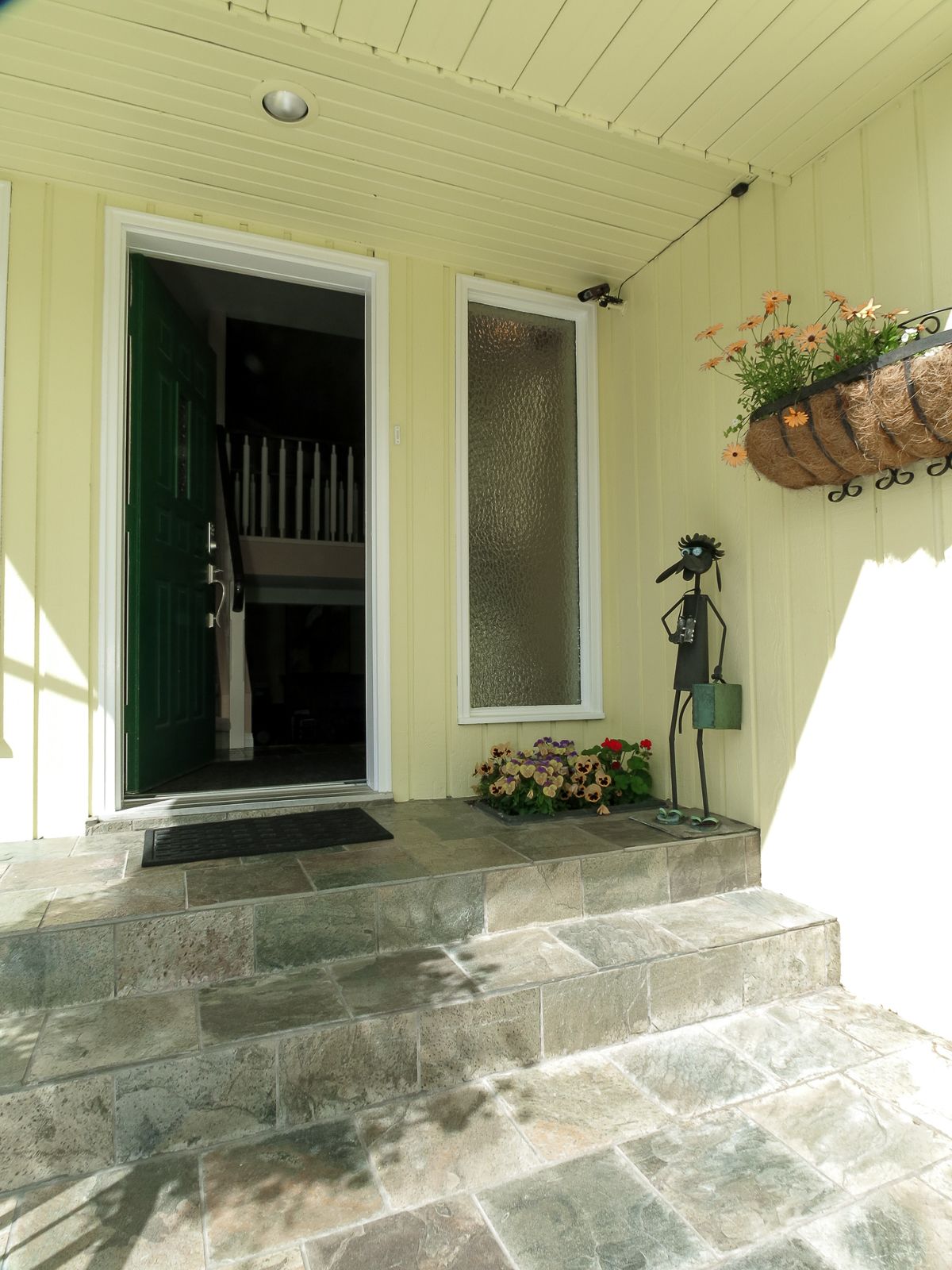 Une entrée de maison avec une porte verte, un petit porche avec des marches, une fenêtre à vitres dépolies et un jardinier à fleurs sur le côté droit.