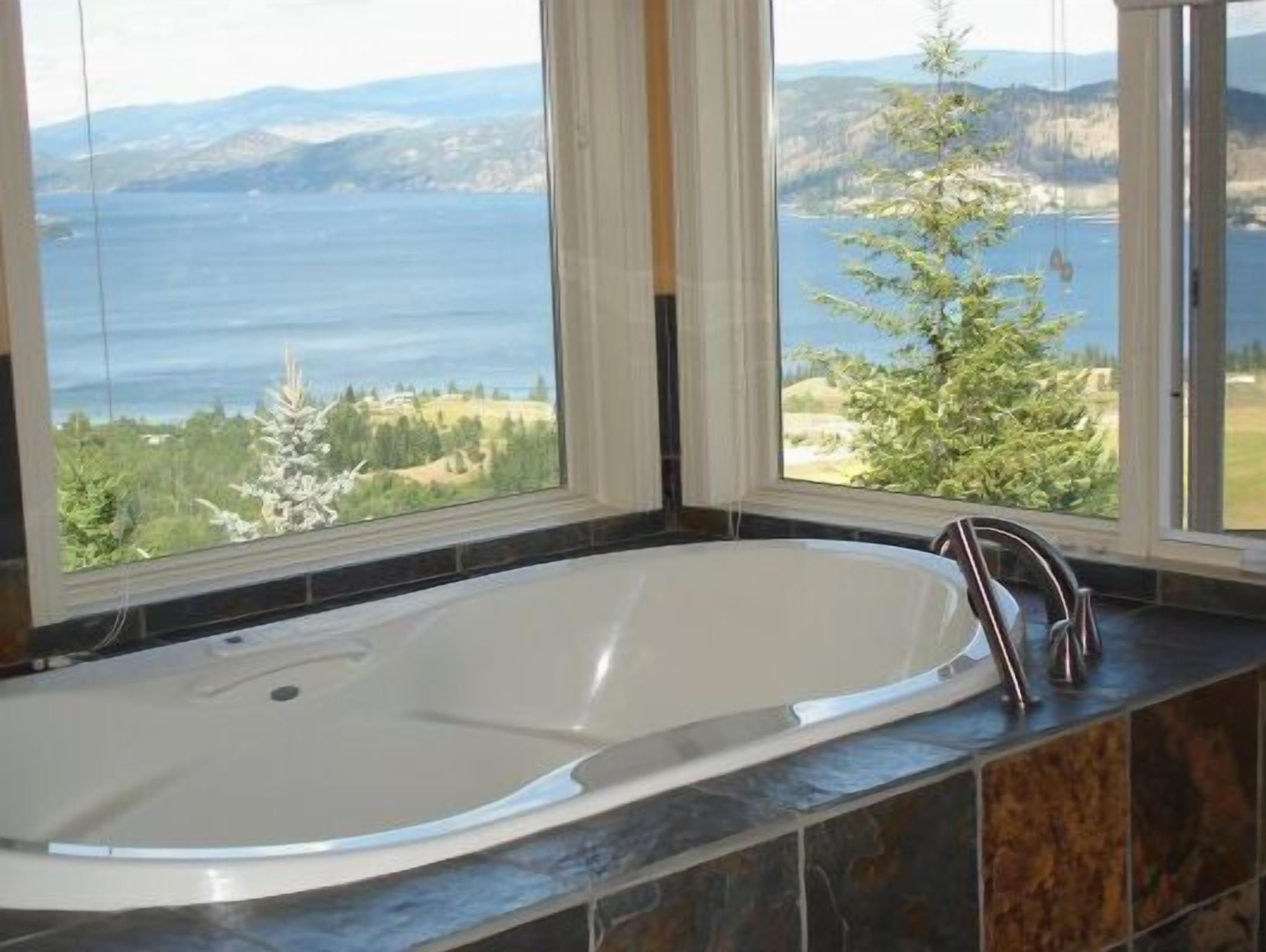 Une salle de bain dotée d’une grande baignoire, offrant une vue sur un lac et des montagnes, et comprenant une fenêtre avec des rideaux.