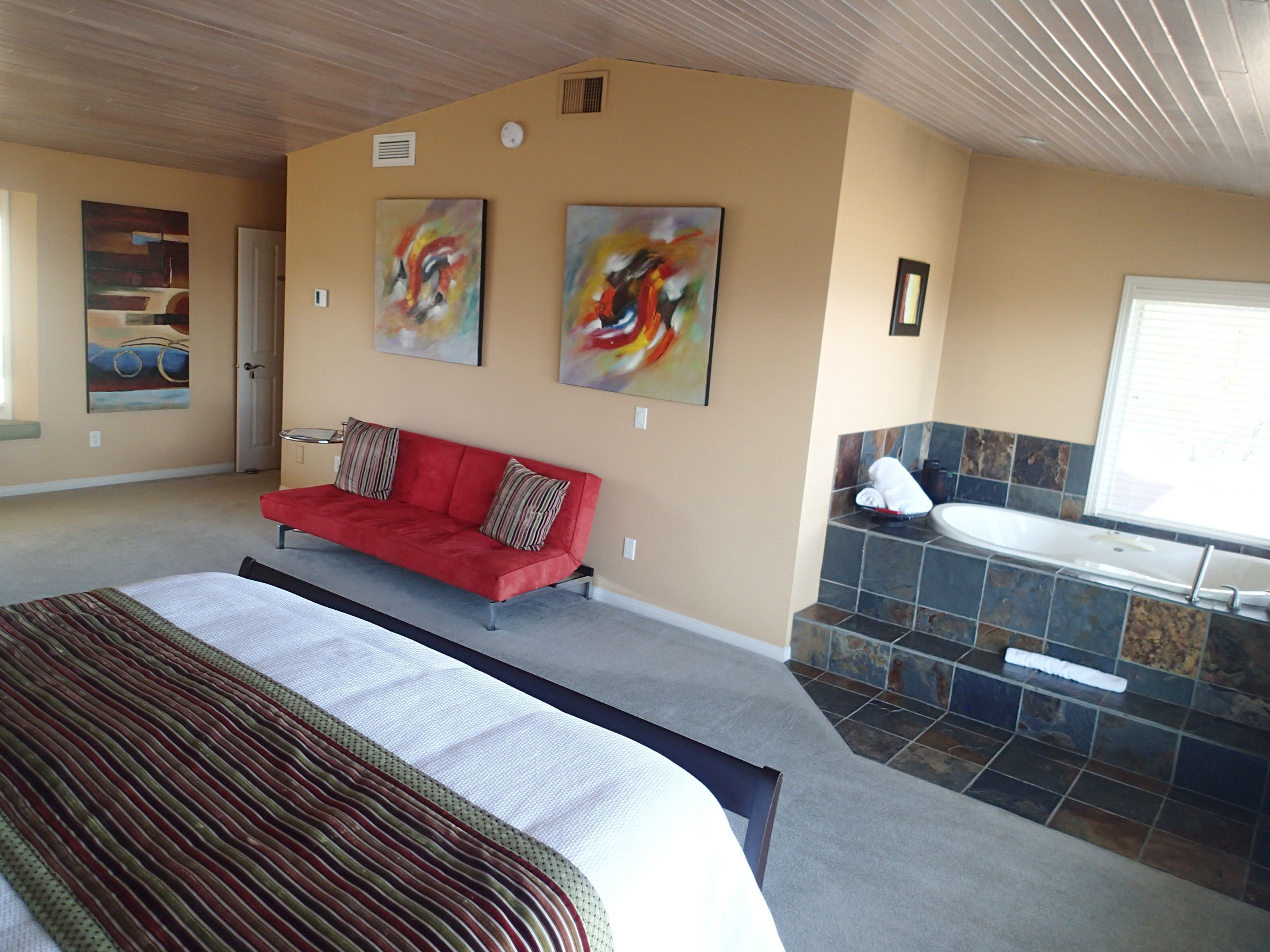 Une chambre avec un grand lit, un canapé rouge, deux tableaux abstraits, une salle de bain avec une baignoire et un espace de douche carrelé.