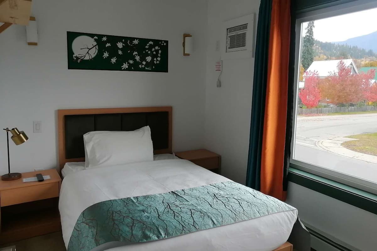 A hotel room with a bed, a nightstand, a lamp, a painting on the wall, an air conditioning unit, and a window with a view of trees.