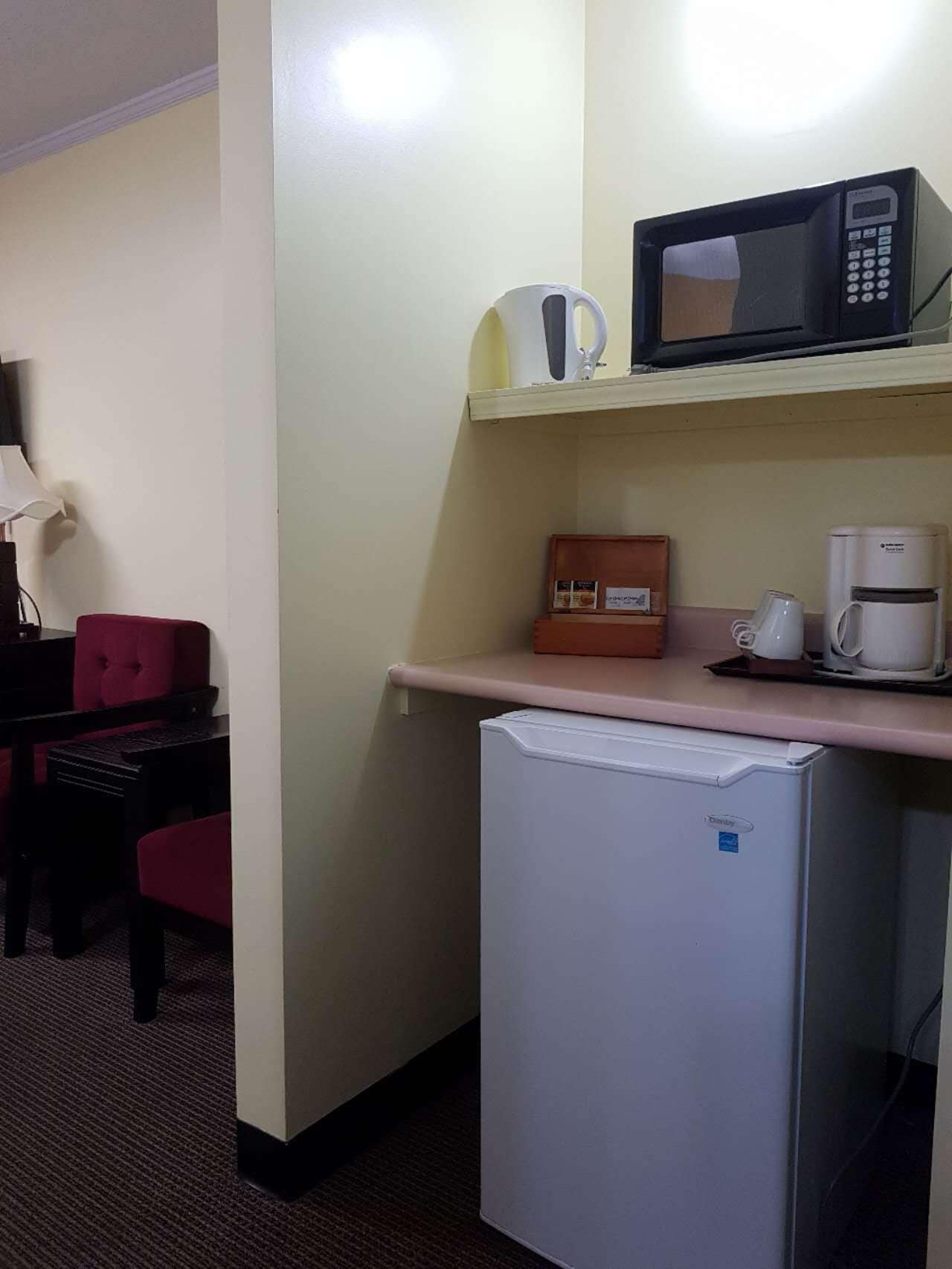 A compact hotel room with a kitchenette including a microwave, coffee maker, and refrigerator. A red chair and a small table are visible in the corner.
