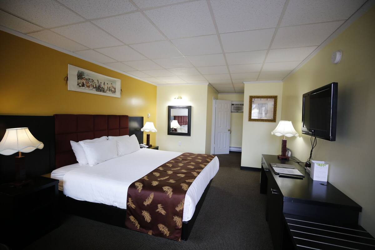 A hotel room with a large bed, two bedside lamps, a flat-screen TV, a desk with a lamp, and a door leading to another room.