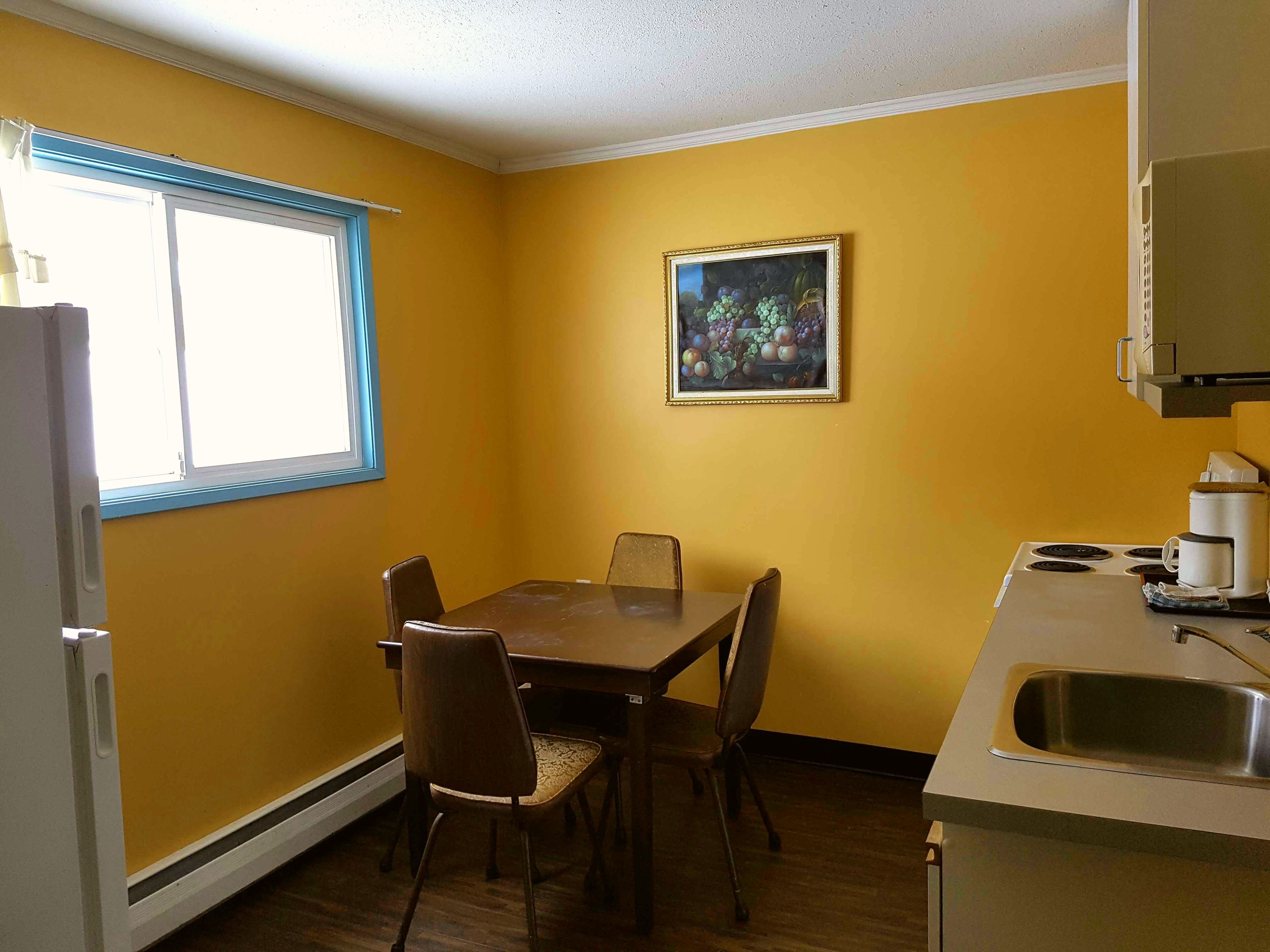 Une cuisine de taille réduite comprenant une table et des chaises, une fenêtre avec des stores, un tableau accroché au mur et un évier.