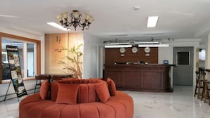 Un hall d’hôtel moderne avec un canapé rond orange, un comptoir d’accueil, un lustre et une grande fenêtre.