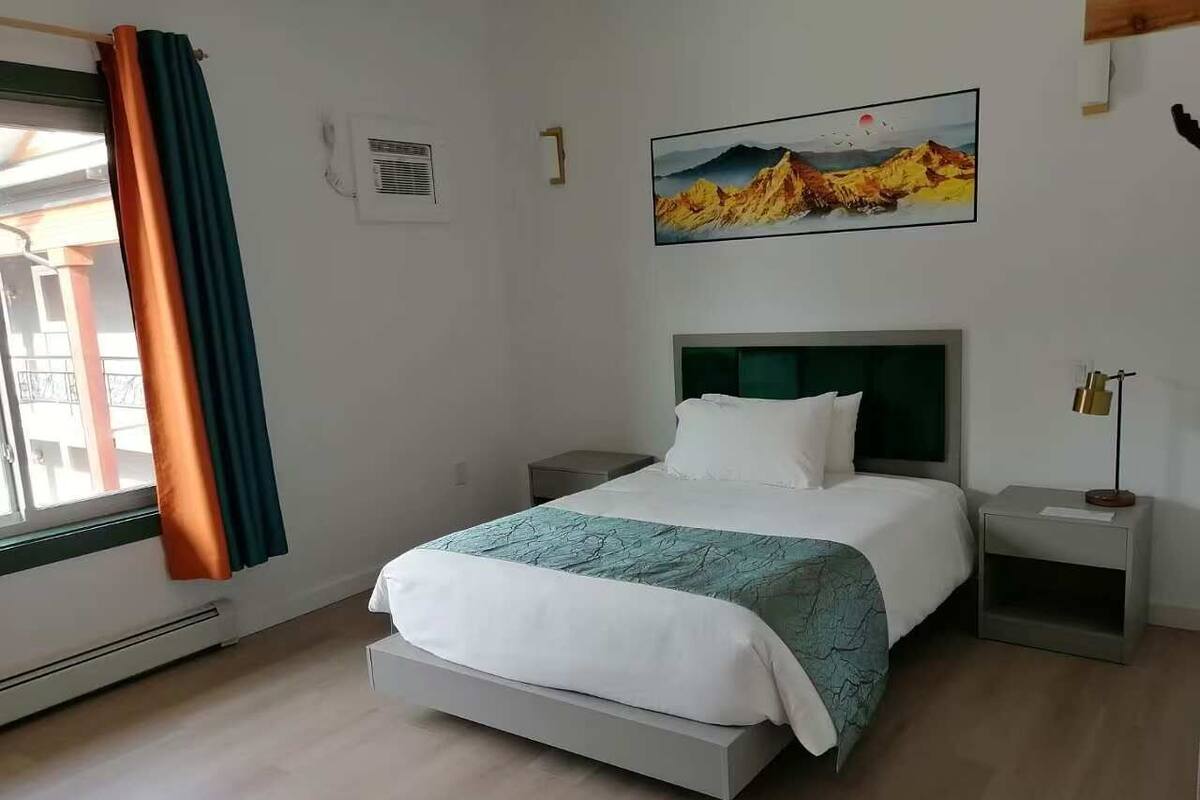A bedroom with a bed, nightstands, a window with curtains, a wall-mounted air conditioner, and a painting.
