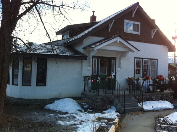 A two-story house with a stone foundation, a front porch with a railing, and a small garden area with snow.