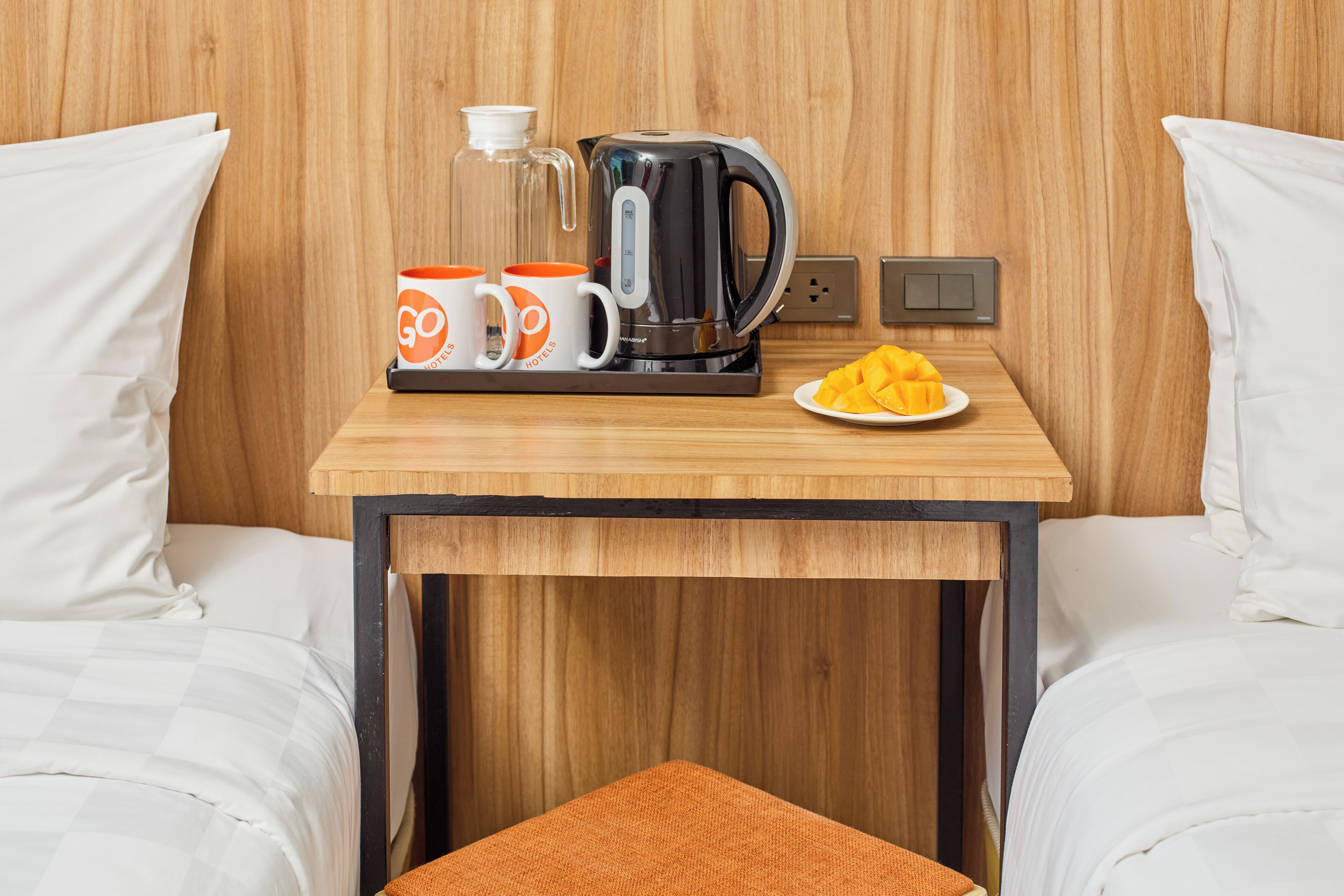 A hotel room with a wooden table, a coffee maker, two mugs, and a plate of fruit.