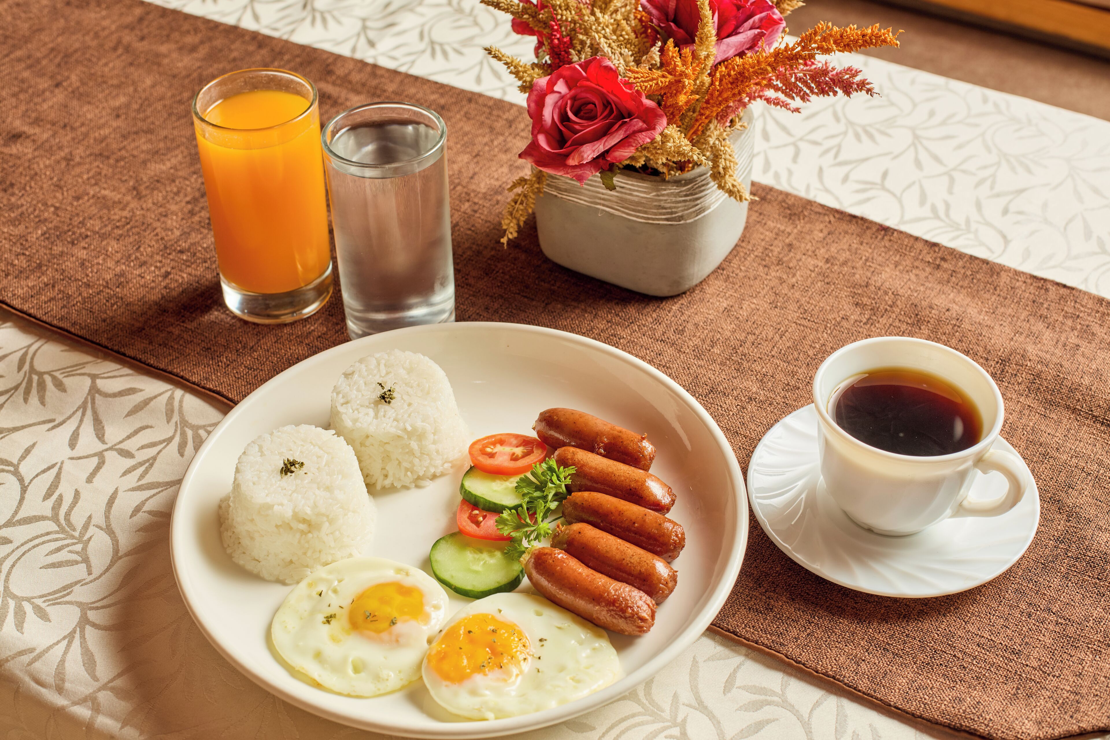 A plate with rice, sausages, eggs, and vegetables, a glass of orange juice, and a cup of coffee on a table with a placemat.