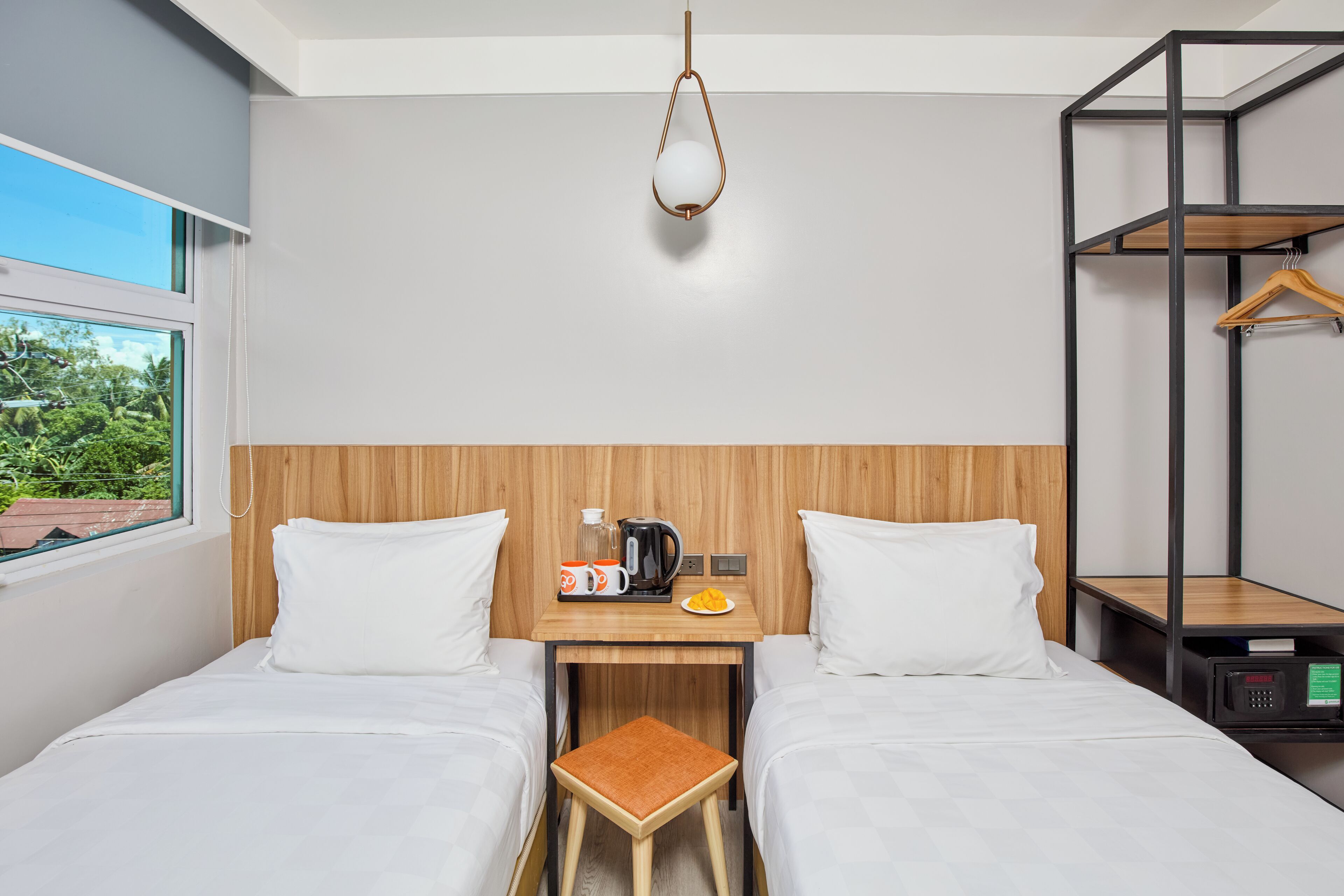 Une chambre d’hôtel avec deux lits, une tête de lit en bois, une petite table avec une machine à café, un luminaire suspendu et une vue sur la verdure.