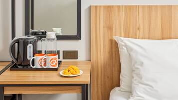 A hotel room with a wooden headboard, a bed with white pillows, a wooden table with a coffee maker, two orange mugs, and a plate of fruit.