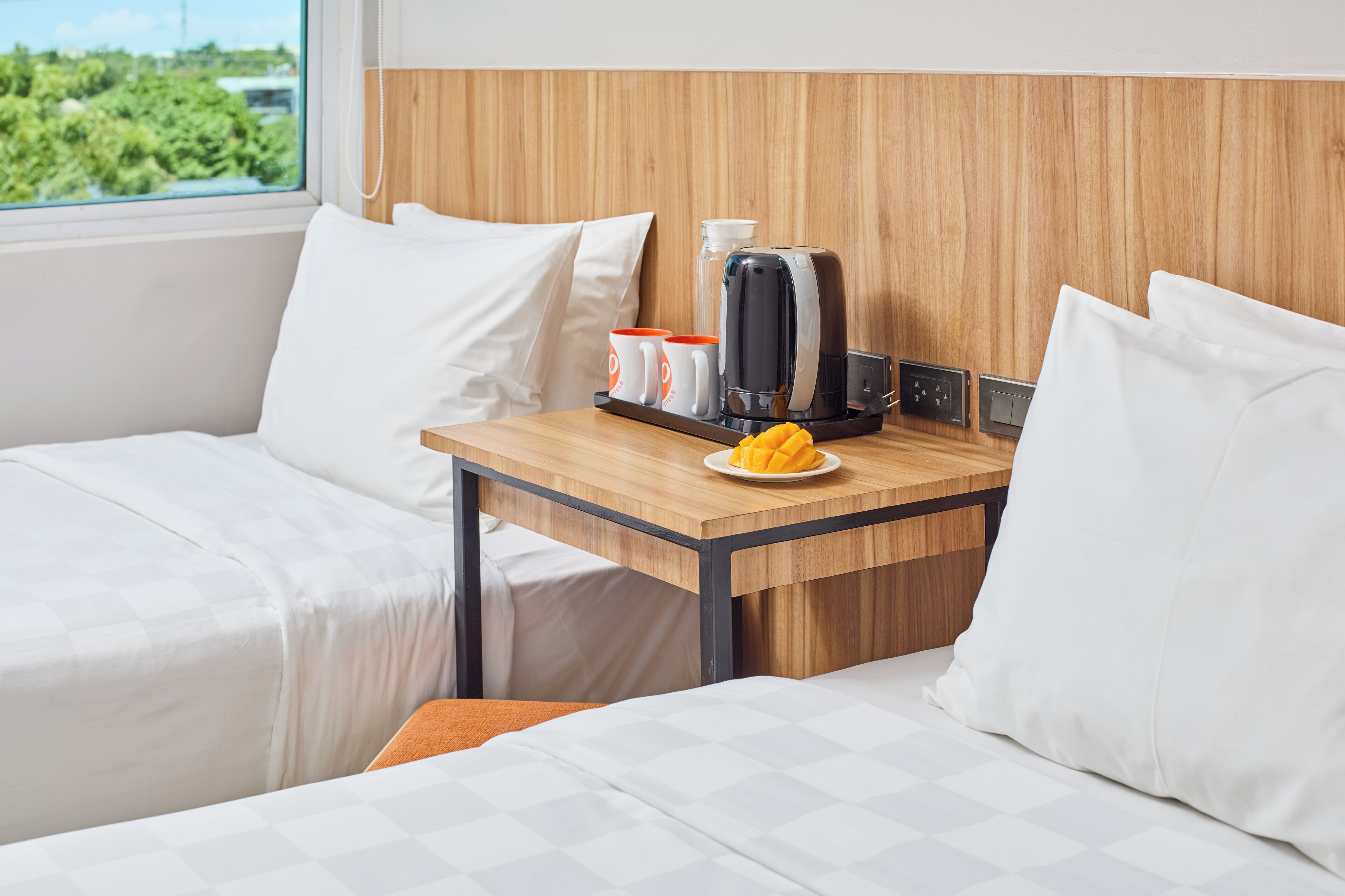 A hotel room with two beds, a wooden headboard, a small table with a coffee maker, cups, and a plate of fruit.