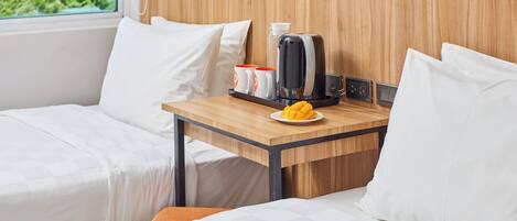 A hotel room with two beds, a wooden headboard, a small table with a coffee maker, cups, and a plate of fruit.