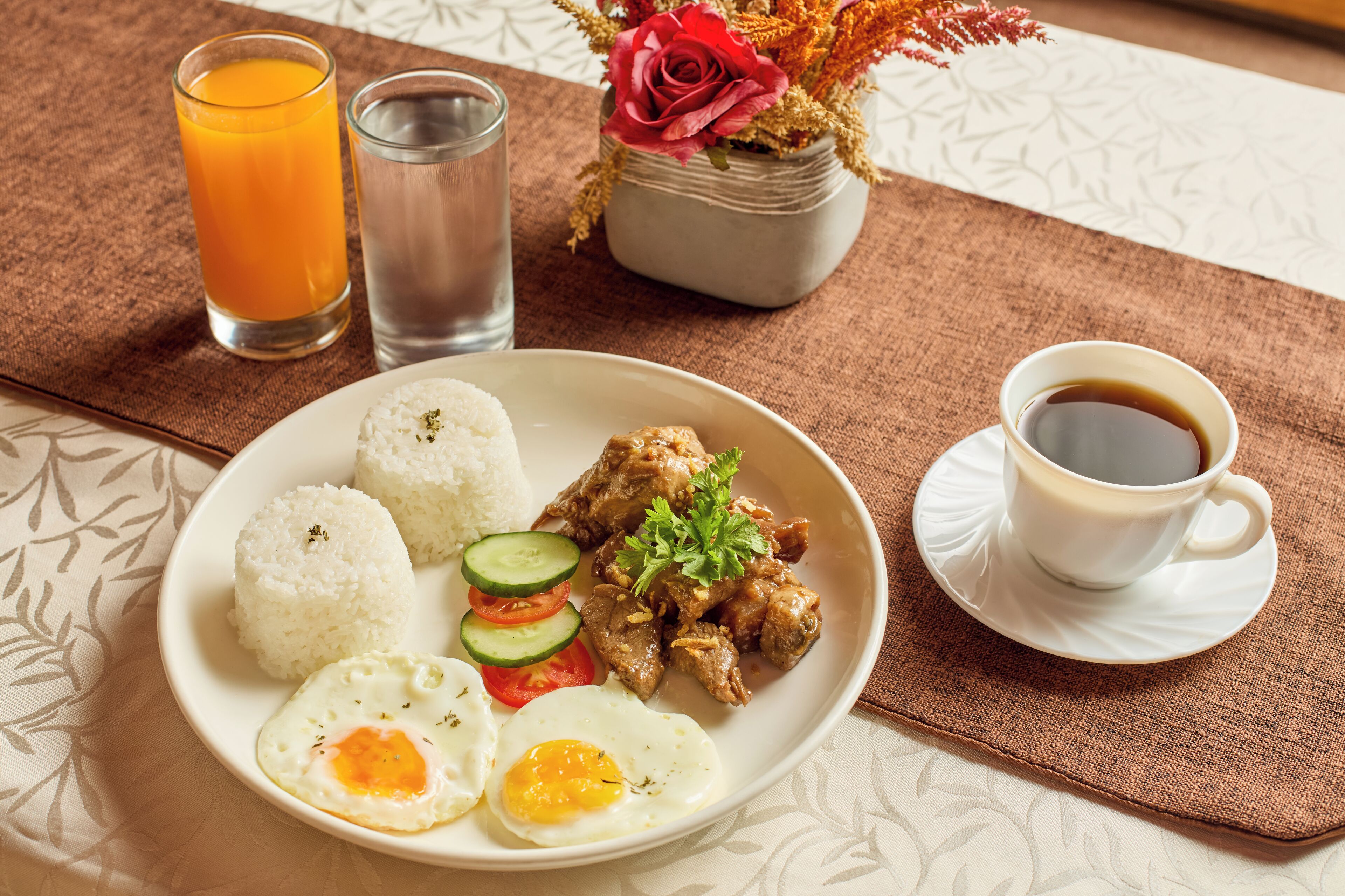 A plate with rice, meat, eggs, and vegetables, a glass of orange juice, a glass of water, and a cup of coffee on a table.