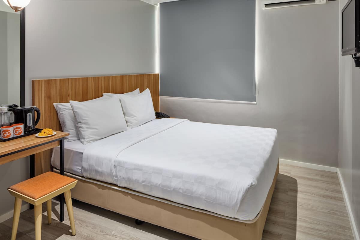 A modern hotel room with a wooden headboard, a bed with white bedding, a small wooden stool, a desk with a coffee maker, and a wall-mounted TV.