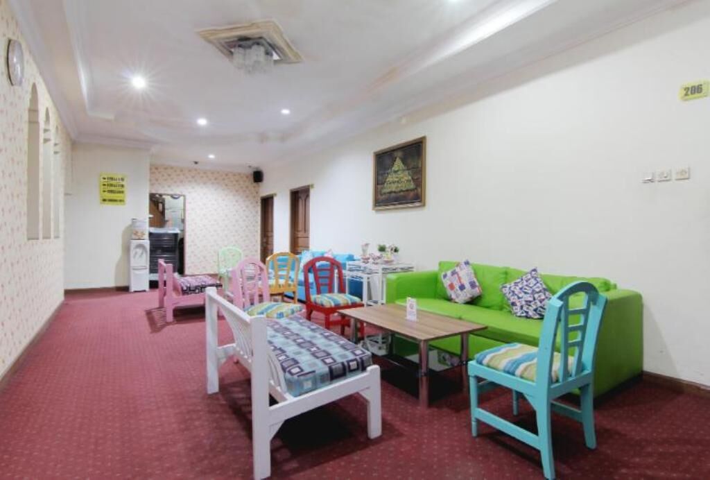 Una habitación con un sofá verde, una mesa de madera y varias sillas con cojines de colores. Hay una pequeña mesa con una lámpara y algunos objetos decorativos encima. La habitación tiene una alfombra roja y paredes blancas con un papel tapiz estampado en una pared.