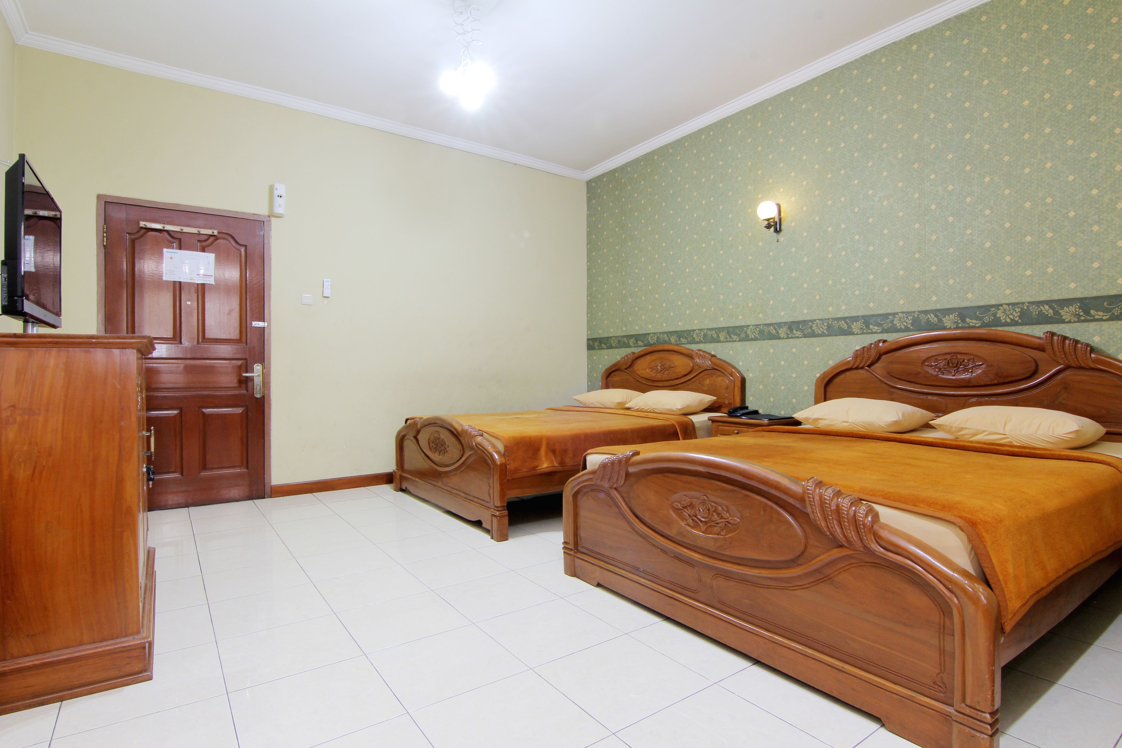 A hotel room with two wooden beds, a wooden wardrobe, a door, and a light fixture on the wall.