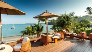 A wooden deck with wicker furniture and umbrellas overlooking a beach with boats and palm trees. - Bandara Pool Villas, Phuket (Wichit)