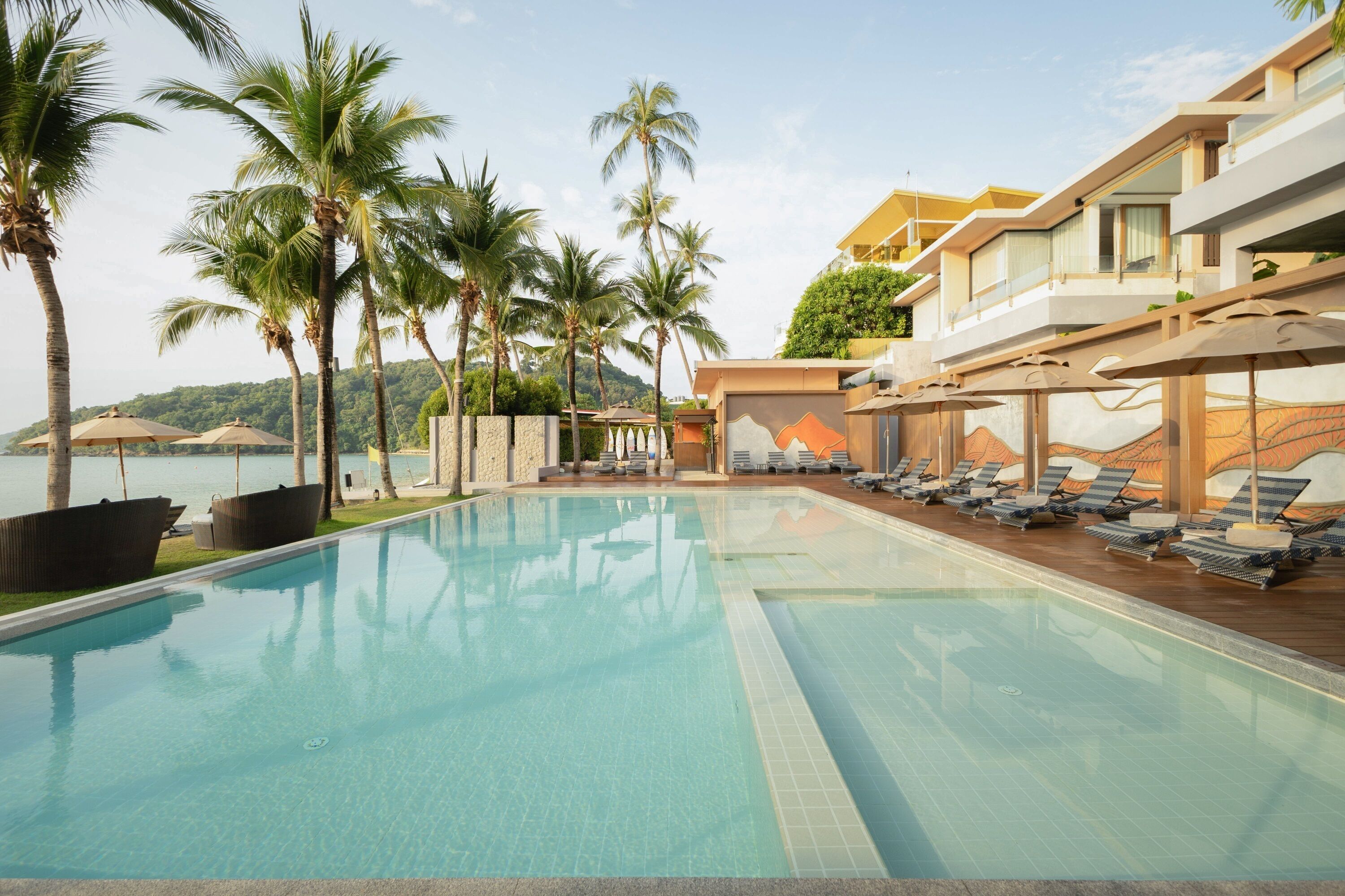Una piscina con sillas de descanso y sombrillas, rodeada de palmeras y vista al mar.
