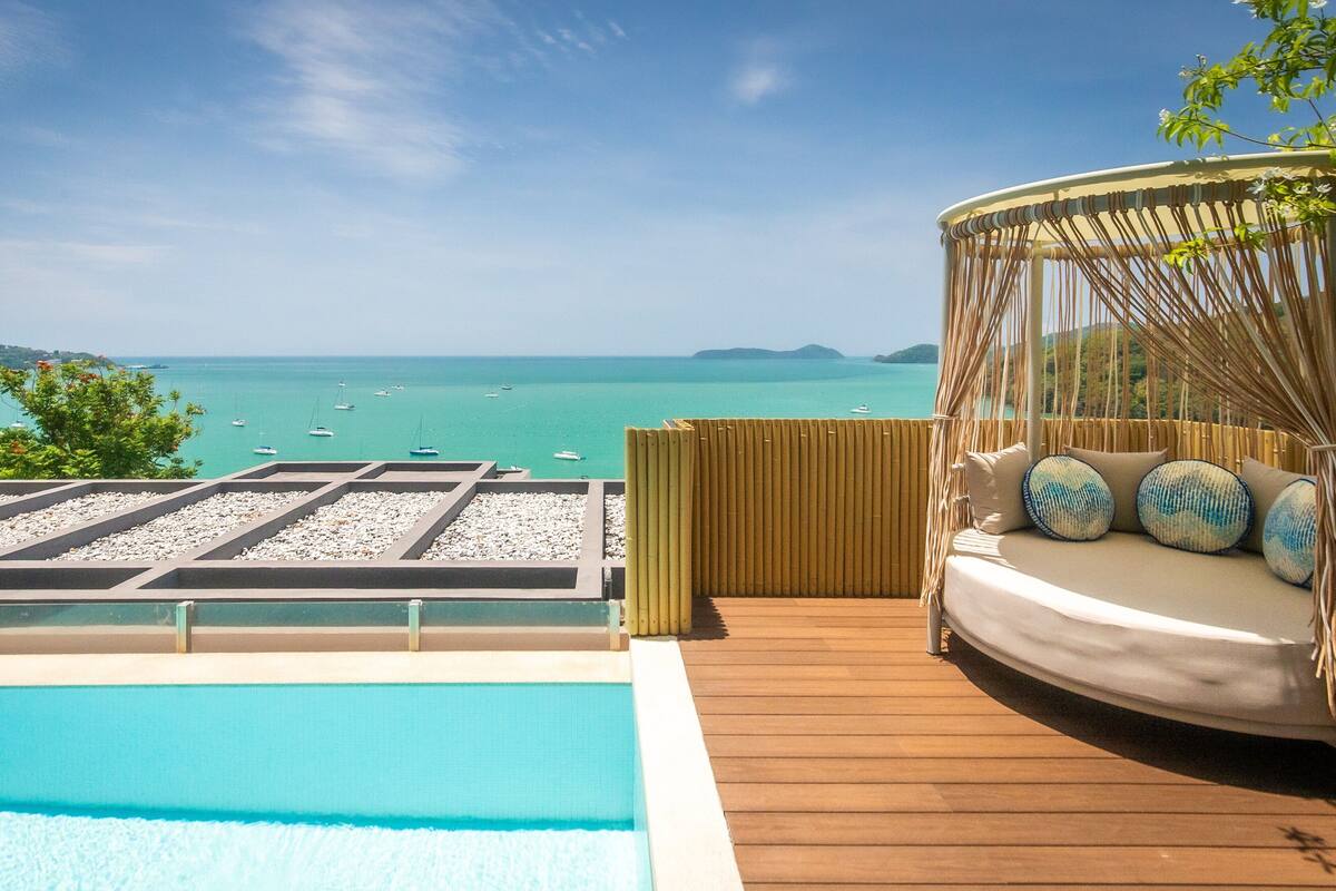 Zona de piscina con terraza de madera, un espacio de descanso circular con cojines y vistas al mar con barcos.