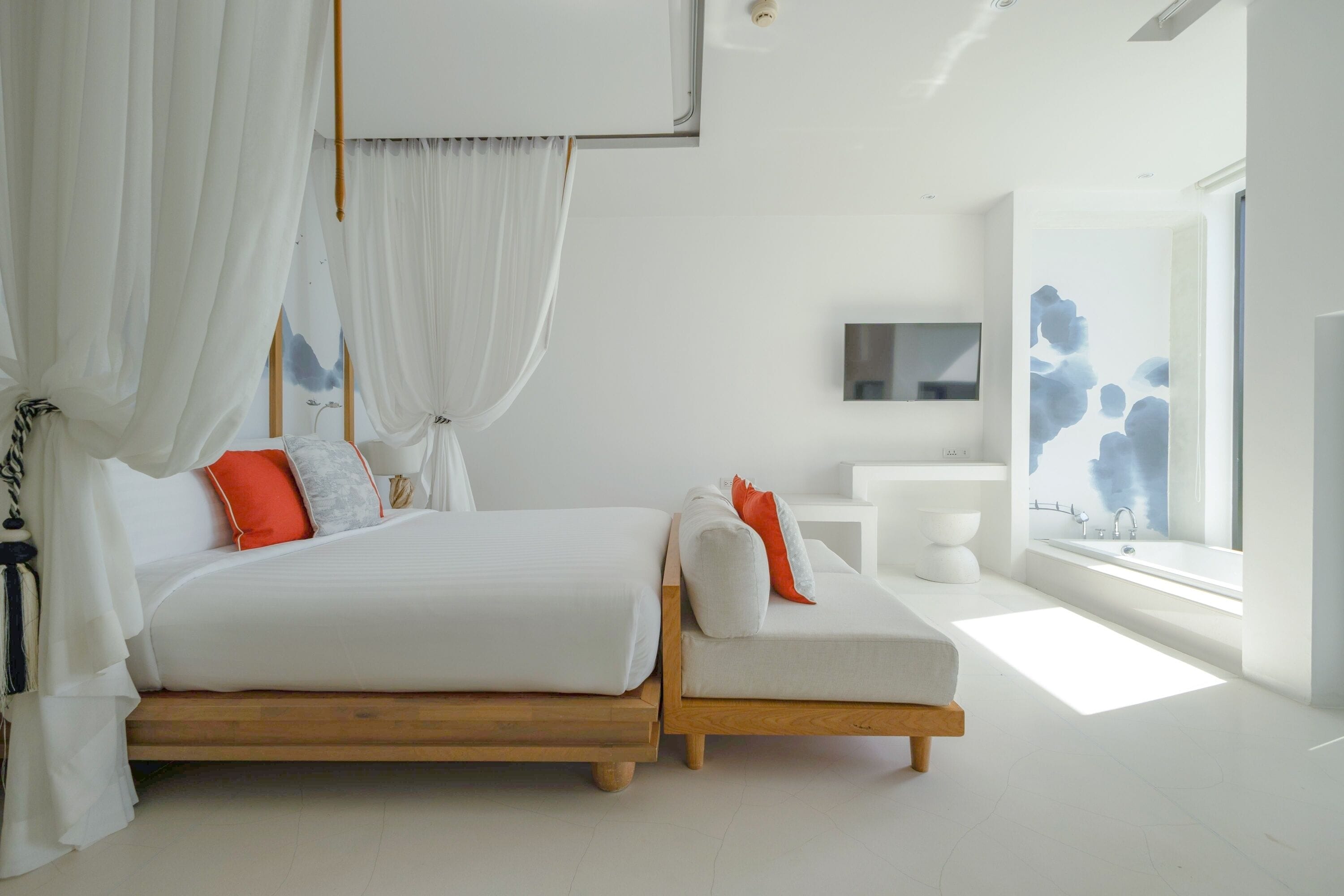 A modern bedroom with a large bed, a daybed, a TV, a bathroom, and a large window with artwork.