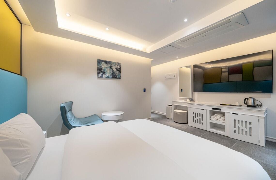 Habitación de hotel moderna con una cama grande, zona de estar con una silla, una mesita redonda, tocador con espejo y un televisor montado en la pared.