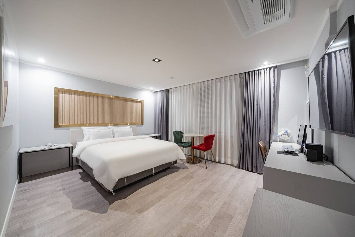 Una camera d'albergo moderna con un letto, comodino, scrivania, sedia e un'ampia finestra con tende.