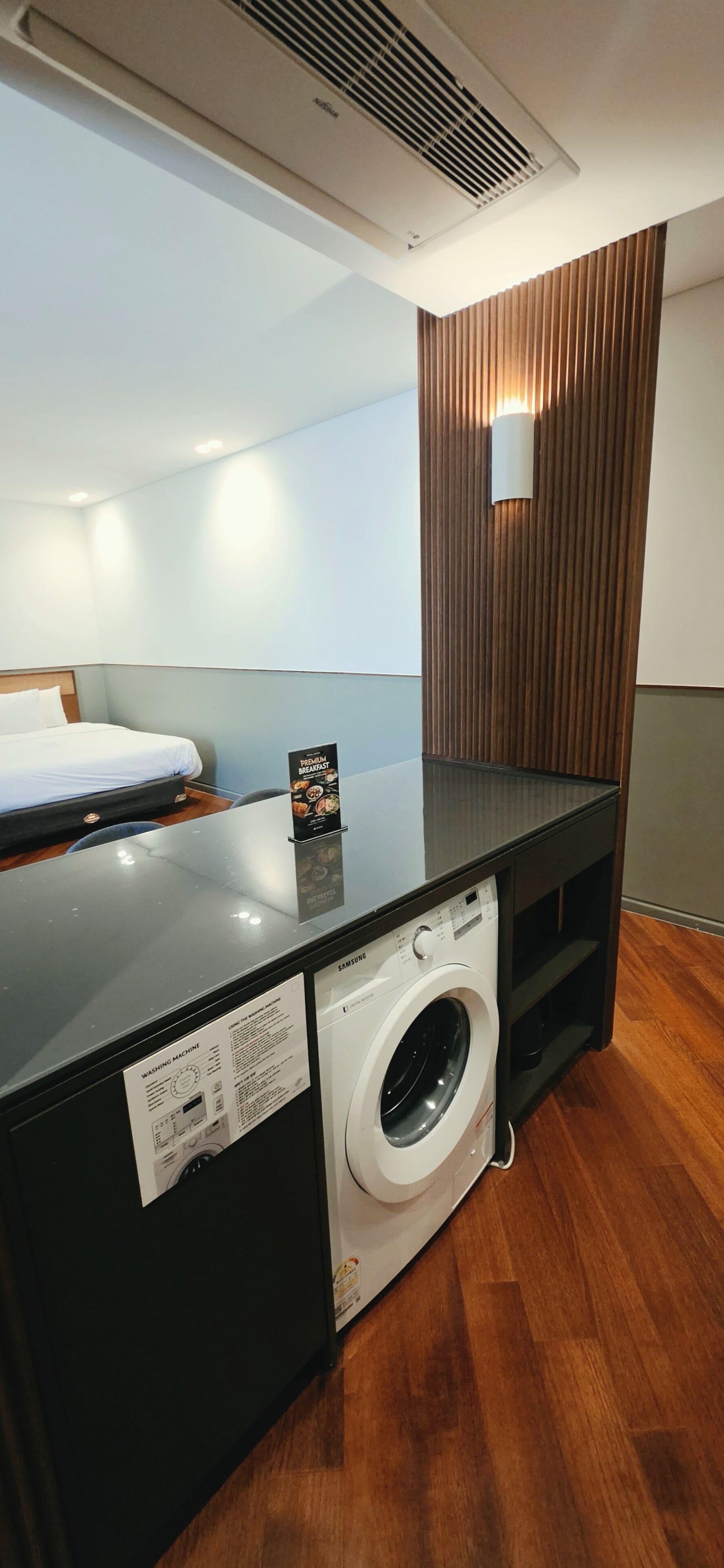 A compact hotel room with a washing machine, a bed, and a wooden panel wall.