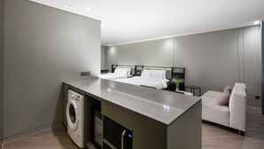 Habitación de hotel moderna con lavadora, cama con ropa blanca, un pequeño rincón de descanso y una kitchenette.