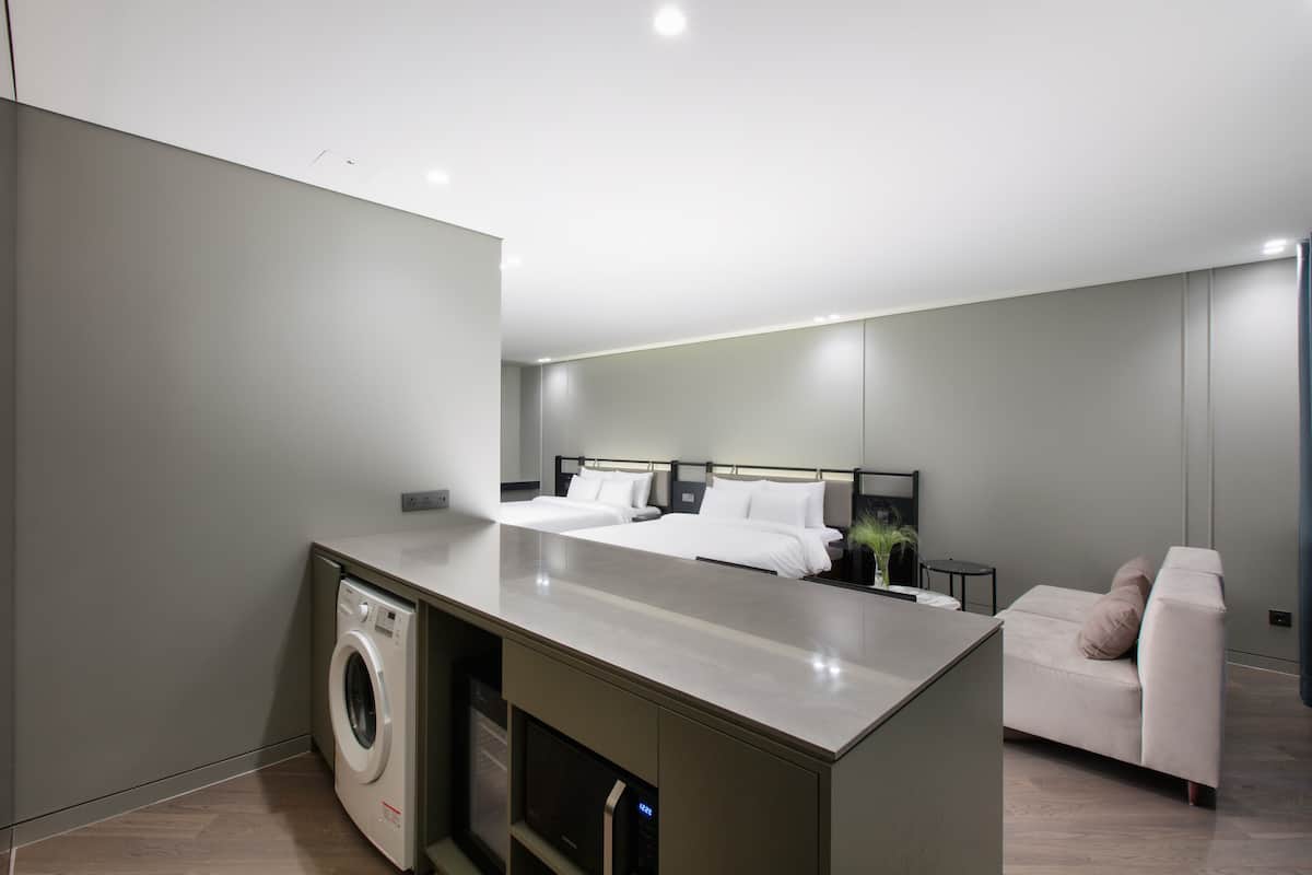 Habitación de hotel moderna con lavadora, cama con ropa blanca, un pequeño rincón de descanso y una kitchenette.